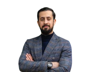 Portrait of Mehmet Yıldız with a short beard, wearing a plaid blazer over a dark turtleneck and a wristwatch, standing with his arms crossed on a transparent background.